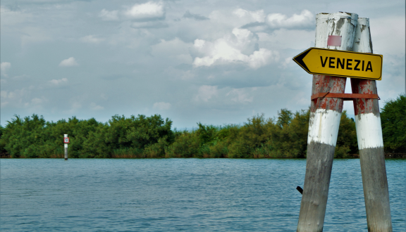 Immagine della laguna di Venezia con isolotto ricoperto di vegetazione e cartello segnaletico giallo su cui si legge "Venezia"